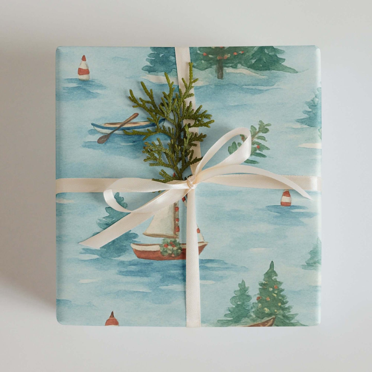 Luxury Christmas wrapping paper with watercolor lake scene and festive sailboat design, topped with natural greenery.