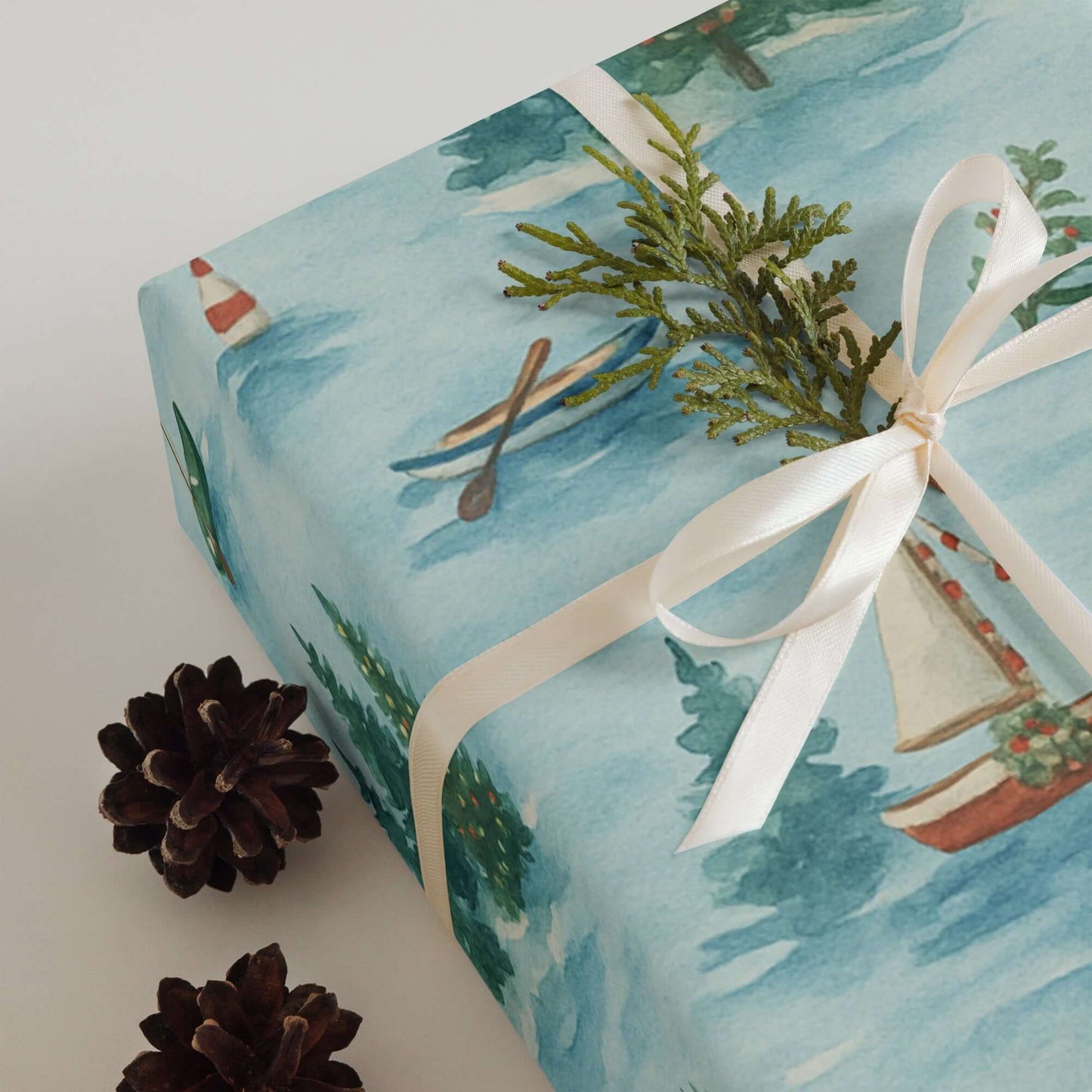 Luxury Christmas wrapping paper featuring hand-painted sailboats and serene snowy lake design, styled with pinecones.