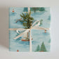 Luxury Christmas wrapping paper with watercolor lake scene and festive sailboat design, topped with natural greenery.