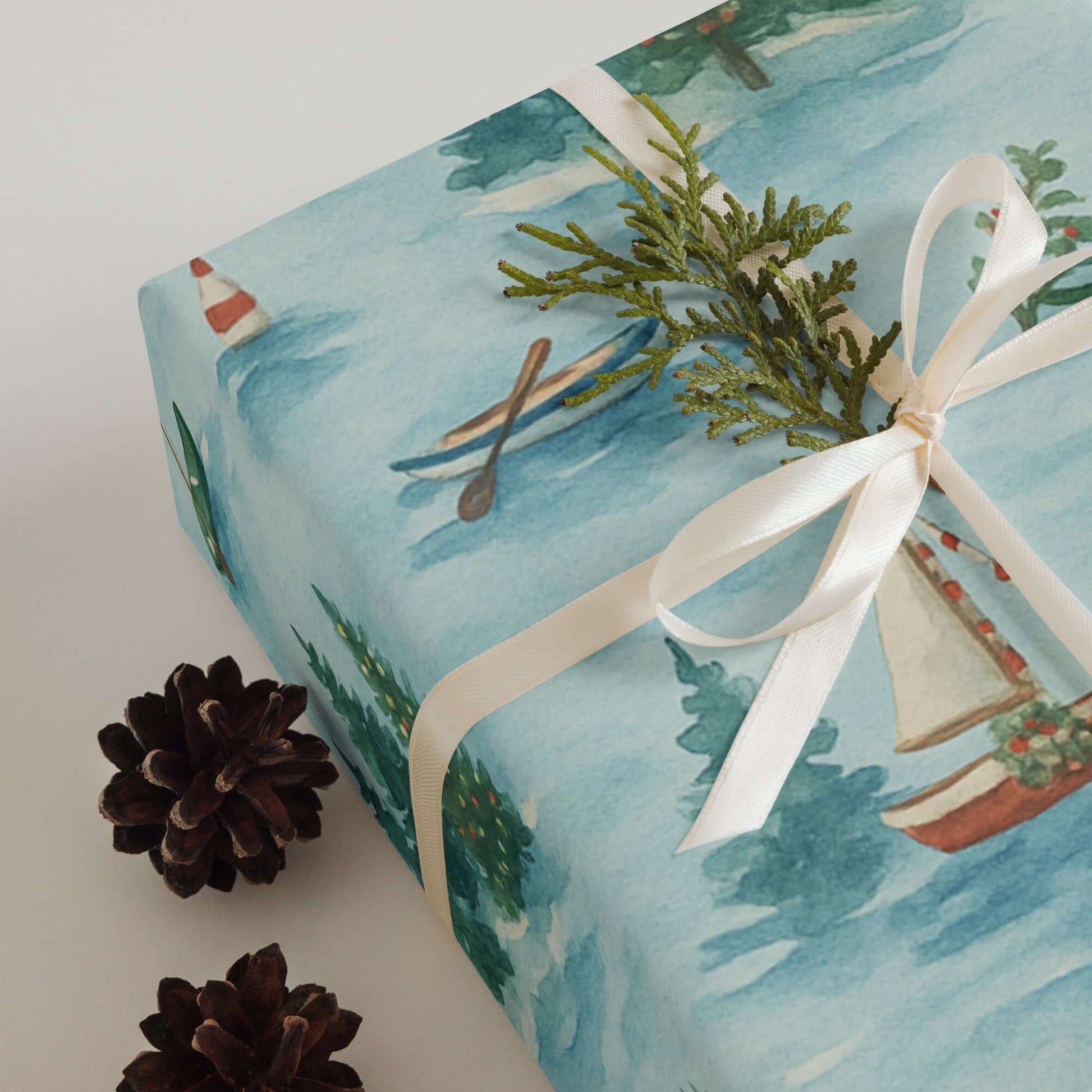 Luxury Christmas wrapping paper featuring hand-painted sailboats and serene snowy lake design, styled with pinecones.