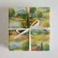Luxury holiday gift wrap featuring a watercolor design of colorful meadows, rustic barns, and distant mountains.