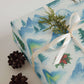 Luxury Christmas wrapping paper featuring watercolor Arctic mountains, northern lights, and holly, styled with pinecones.