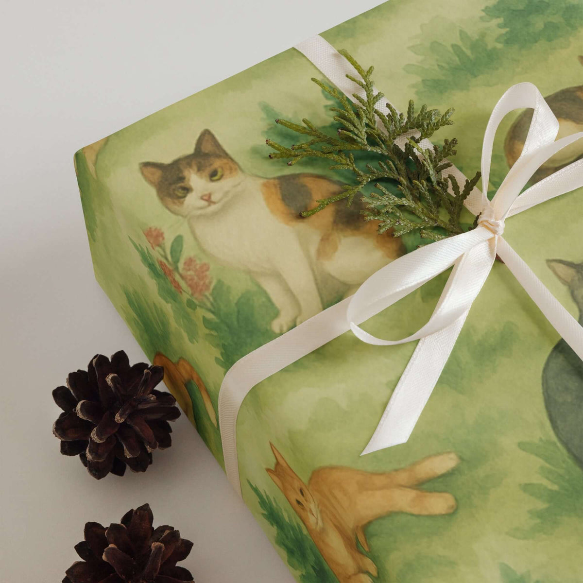 Cats Wrapping Paper featuring adorable cat illustrations among greenery and a decorative bow with pinecones.