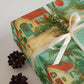 Caribbean Tropical Christmas Wrapping Paper on a gift box adorned with greenery and feminine ribbon, pine cones nearby.